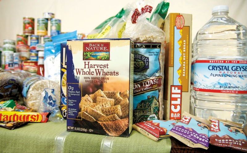 Food sitting on a table, rice cakes, clif bars, cracker, canned goods and water